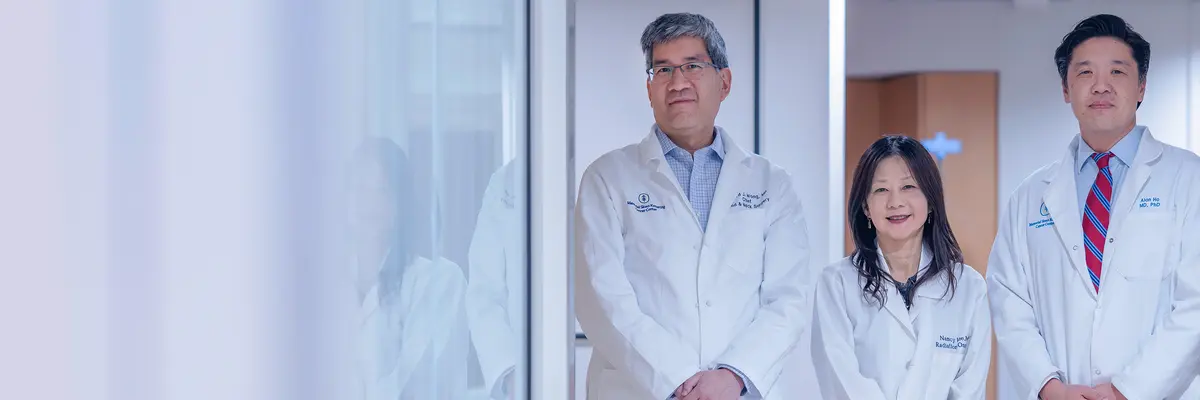 Surgeon Dr. Richard Wong, radiation oncologist Dr. Nancy Lee, and medical oncologist Dr. Alan Ho, wearing white doctors’ coats, stand in a hallway at MSK in NYC.