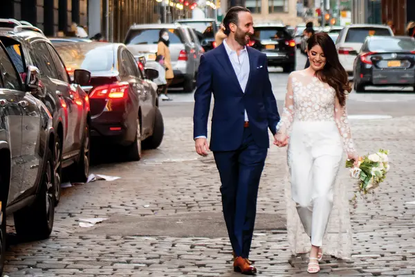 Cancer survivor Rebecca Lerner and her husband Ian smile and walk on a city street, holding hands on their wedding day. 