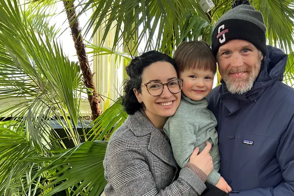 Kevin Tougher and his wife, Maria, holding their son, Theo, surrounded by trees outdoors. 
