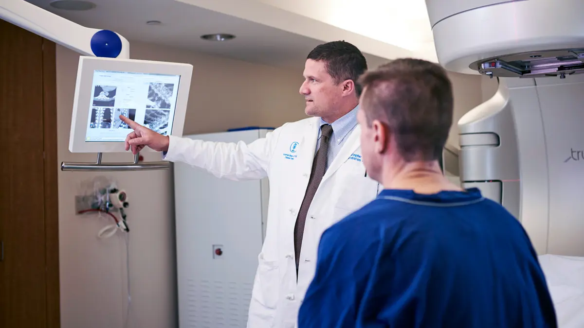 Radiation oncologist Dr. Christopher Crane, pointing at a monitor, with a patient in MSK’s linear accelerator treatment suite. 
