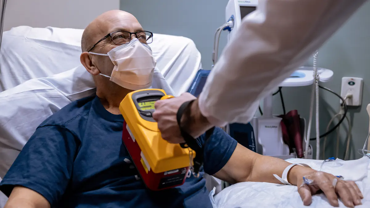 A man wearing a protective mask is in a hospital bed while a healthcare provider holds treatment equipment on his chest.  