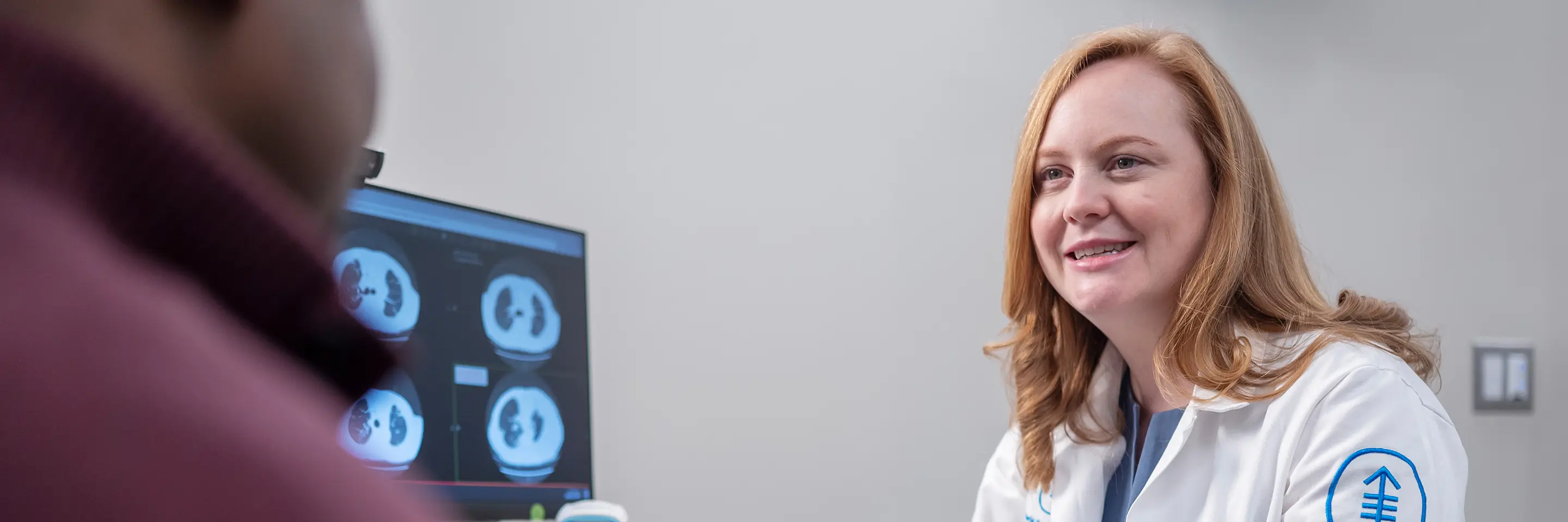 Female doctor in a white coat sitting at a computer speaking with a male patient wearing a maroon shirt.